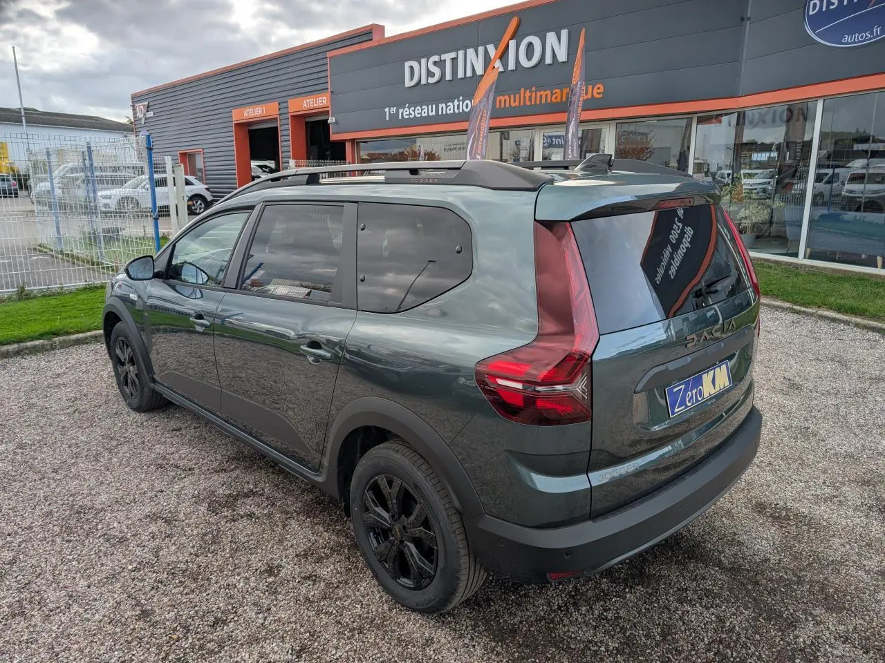 Vue 3/4 arrière droite d'un Dacia Jogger vert métallisé avec jantes noires et barres de toit, stationné devant un garage.