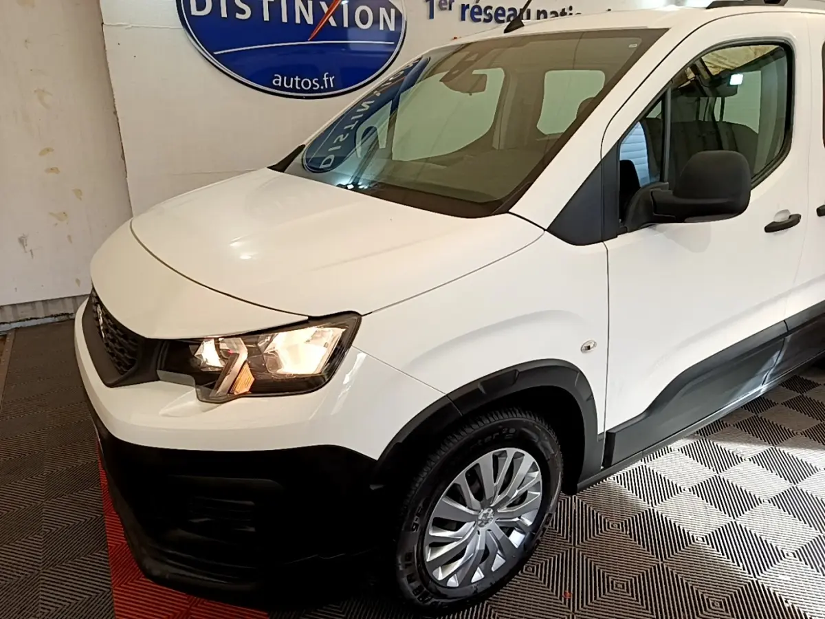 Vue 3/4 avant droit d'un Peugeot Rifter blanc avec phares allumés et protections noires sur les bas de caisse.