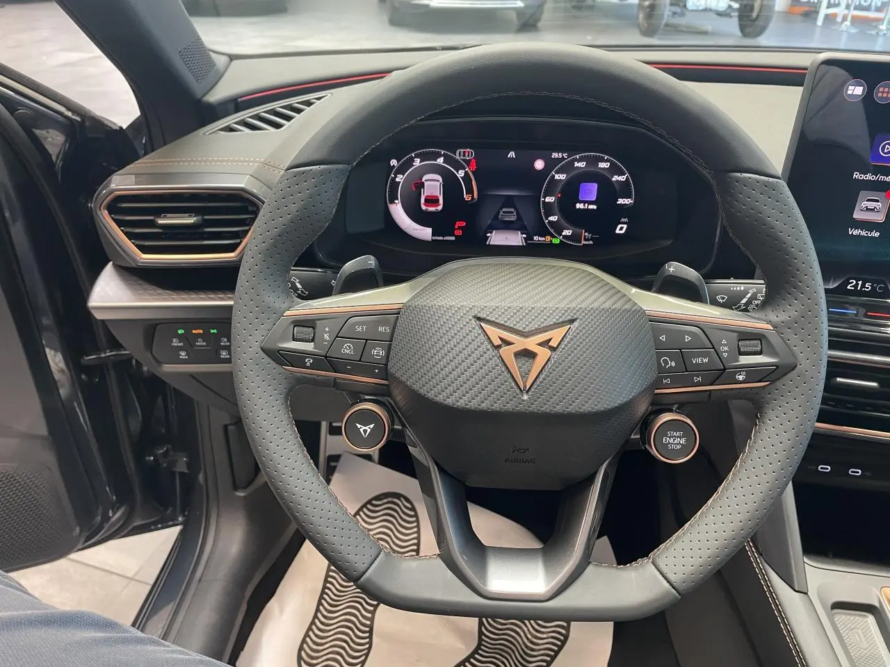 Vue intérieure centrée sur le volant perforé Cupra avec logo cuivré et tableau de bord digital du Leon gris magnetic.