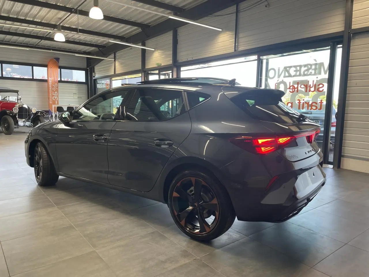 Vue 3/4 arrière droite d'une CUPRA Leon gris magnetic avec jantes cuivrées et feux arrière allumés en intérieur showroom.