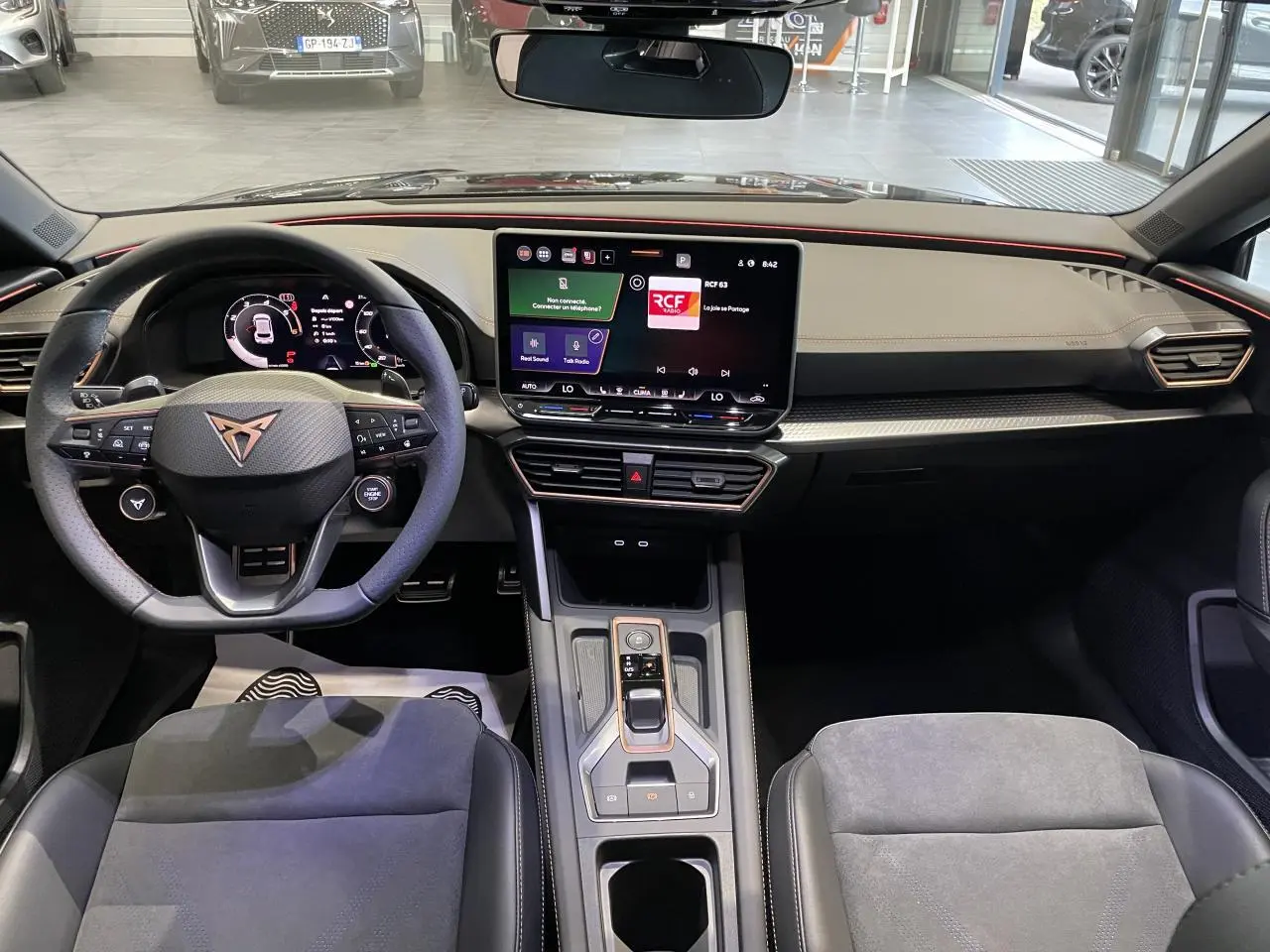 Vue intérieure avant de la CUPRA Leon 2026, tableau de bord noir avec écran tactile central et volant sport à logo cuivré.