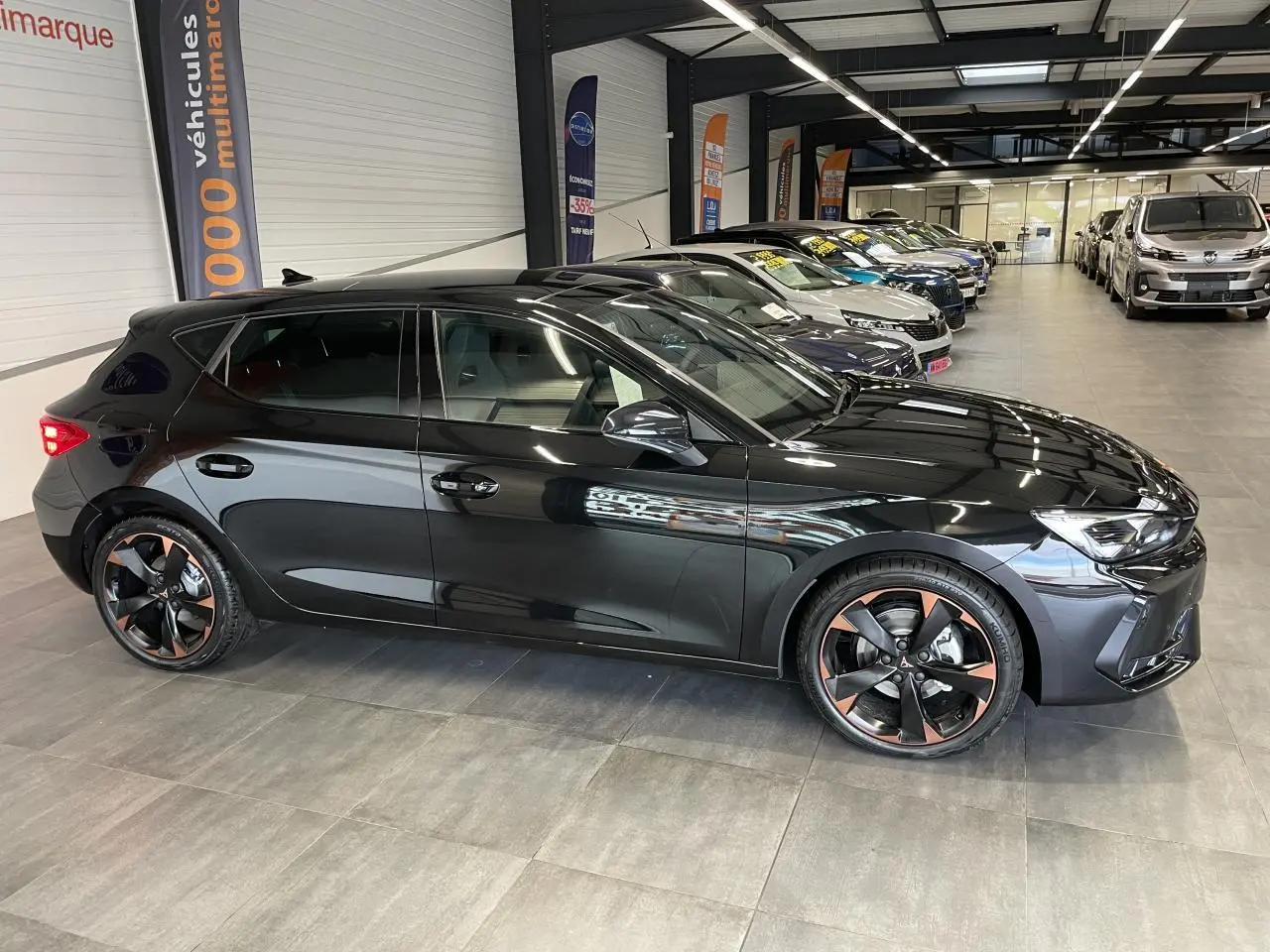 Vue de profil côté gauche d'une CUPRA Leon noire avec jantes alliage 18'' cuivrées dans un showroom.