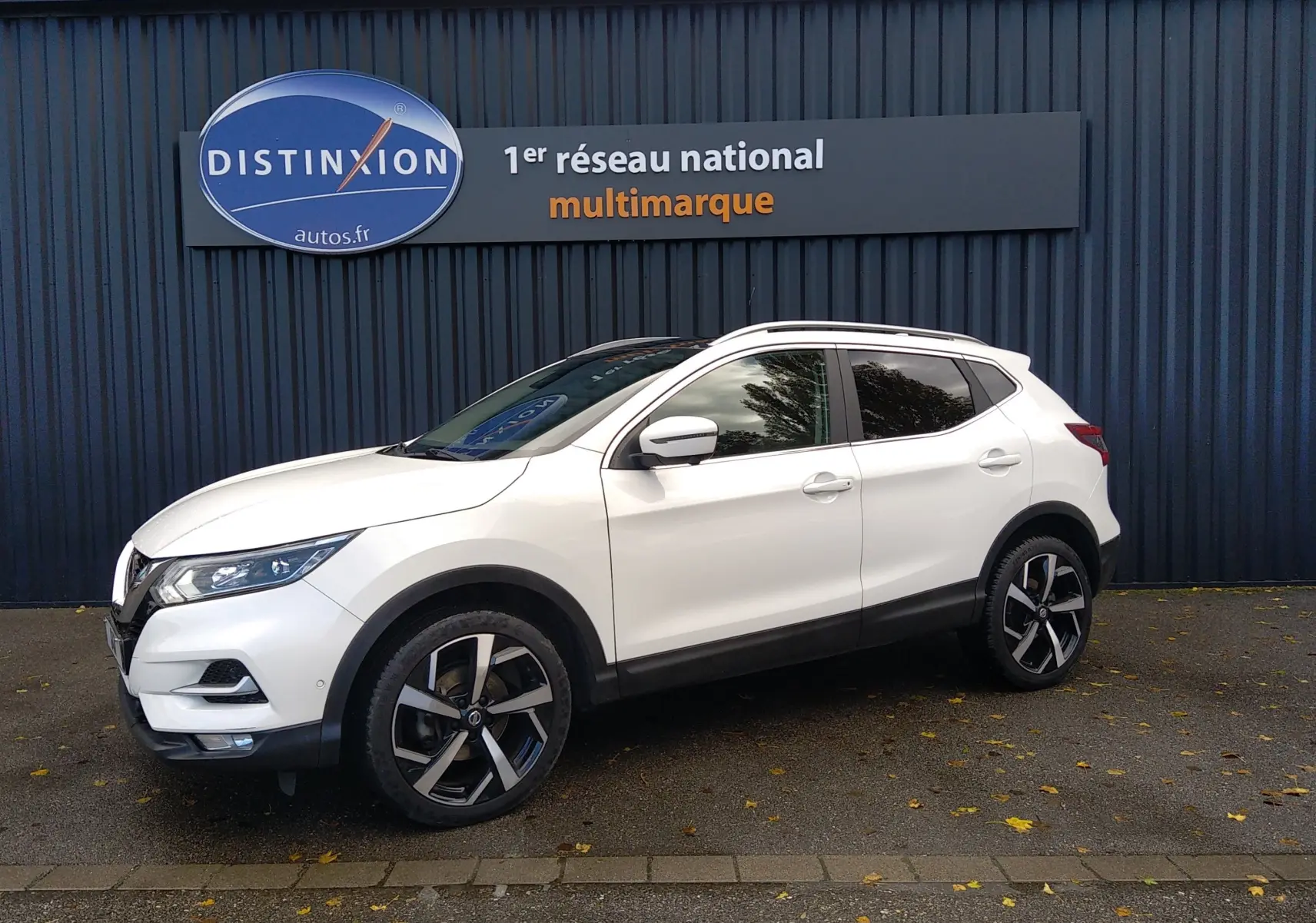 Nissan Qashqai blanc en 3/4 avant droit, avec jantes alliage noires et toit ouvrant, devant un mur sombre.
