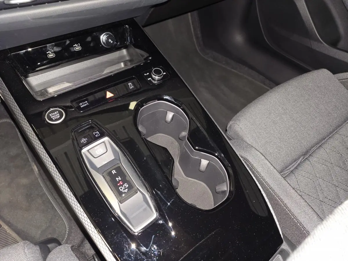 Vue rapprochée de la console centrale noire brillante avec levier de vitesses automatique et porte-gobelets, siège tissu gris visible.