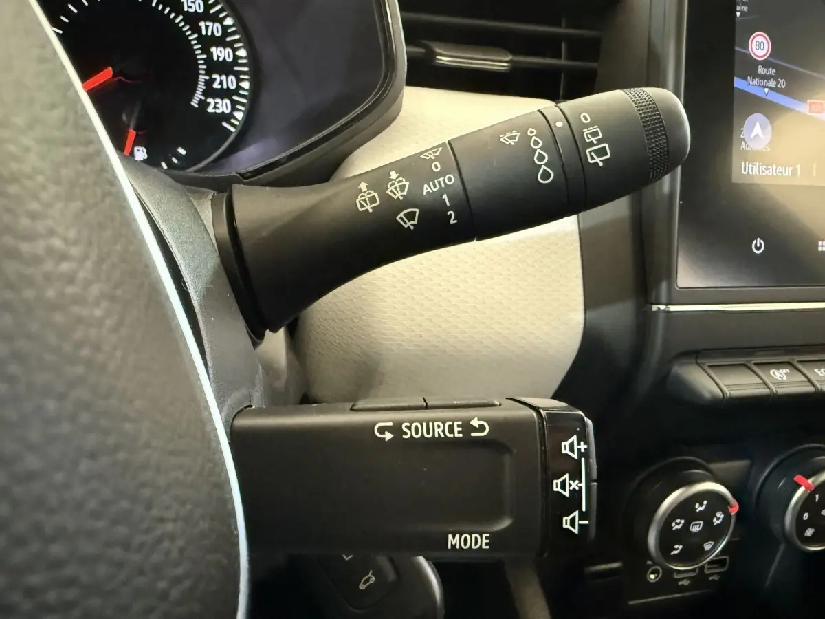 Gros plan sur les commandes au volant et levier d’essuie-glace d’une Renault Clio blanche, tableau de bord partiellement visible.