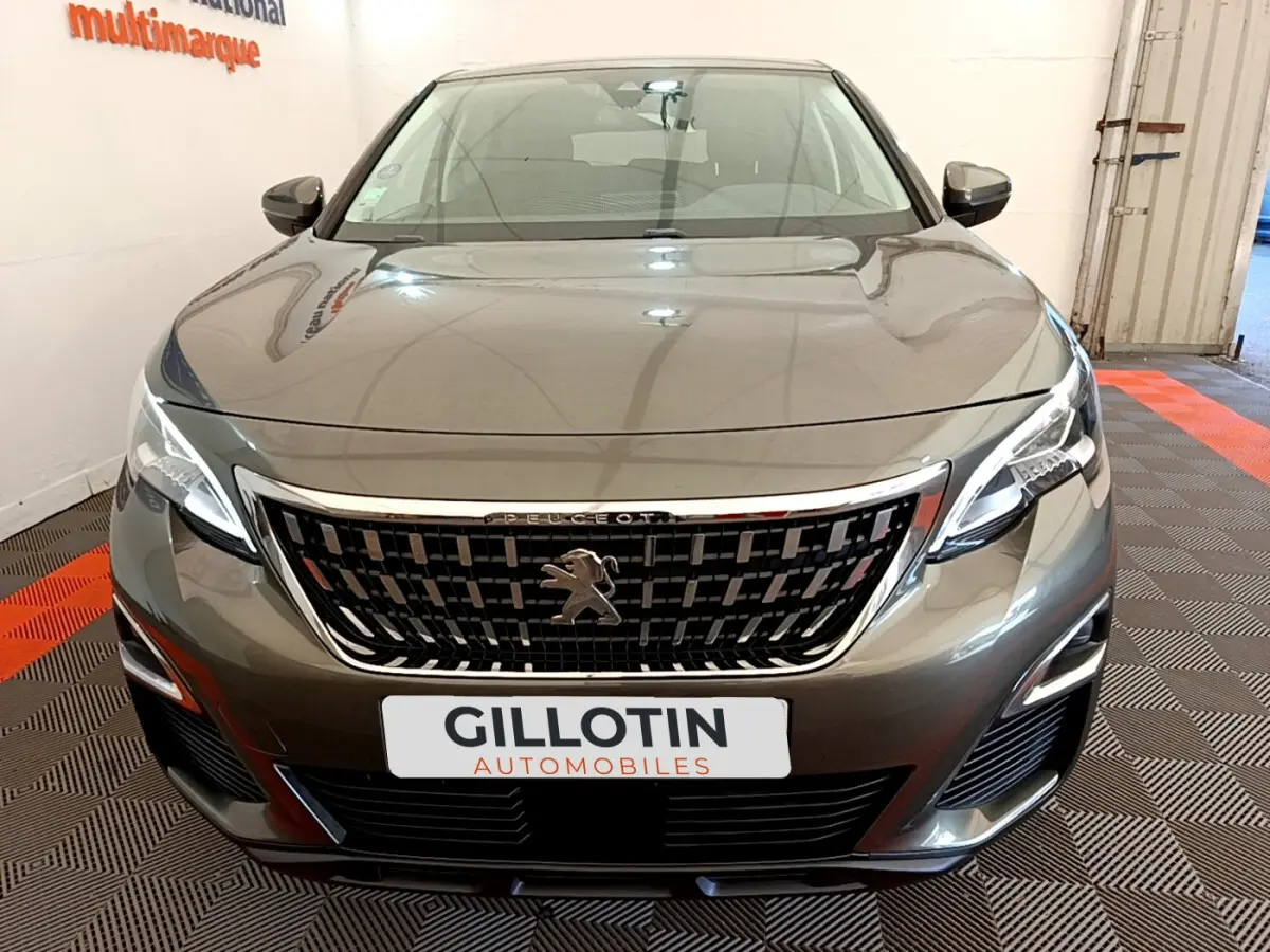 Vue de face d'un Peugeot 3008 gris foncé avec calandre chromée et phares LED allumés en intérieur showroom.