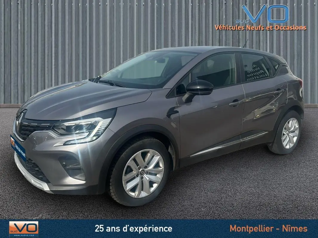 Renault Captur TCe 100 Business gris vu en 3/4 avant droit sur fond industriel, avec feux allumés et jantes argentées.