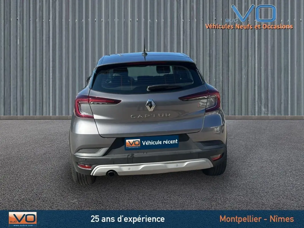 Vue arrière d'un Renault Captur gris 2021 avec feux allumés et plaque "Véhicule récent" sur fond industriel.