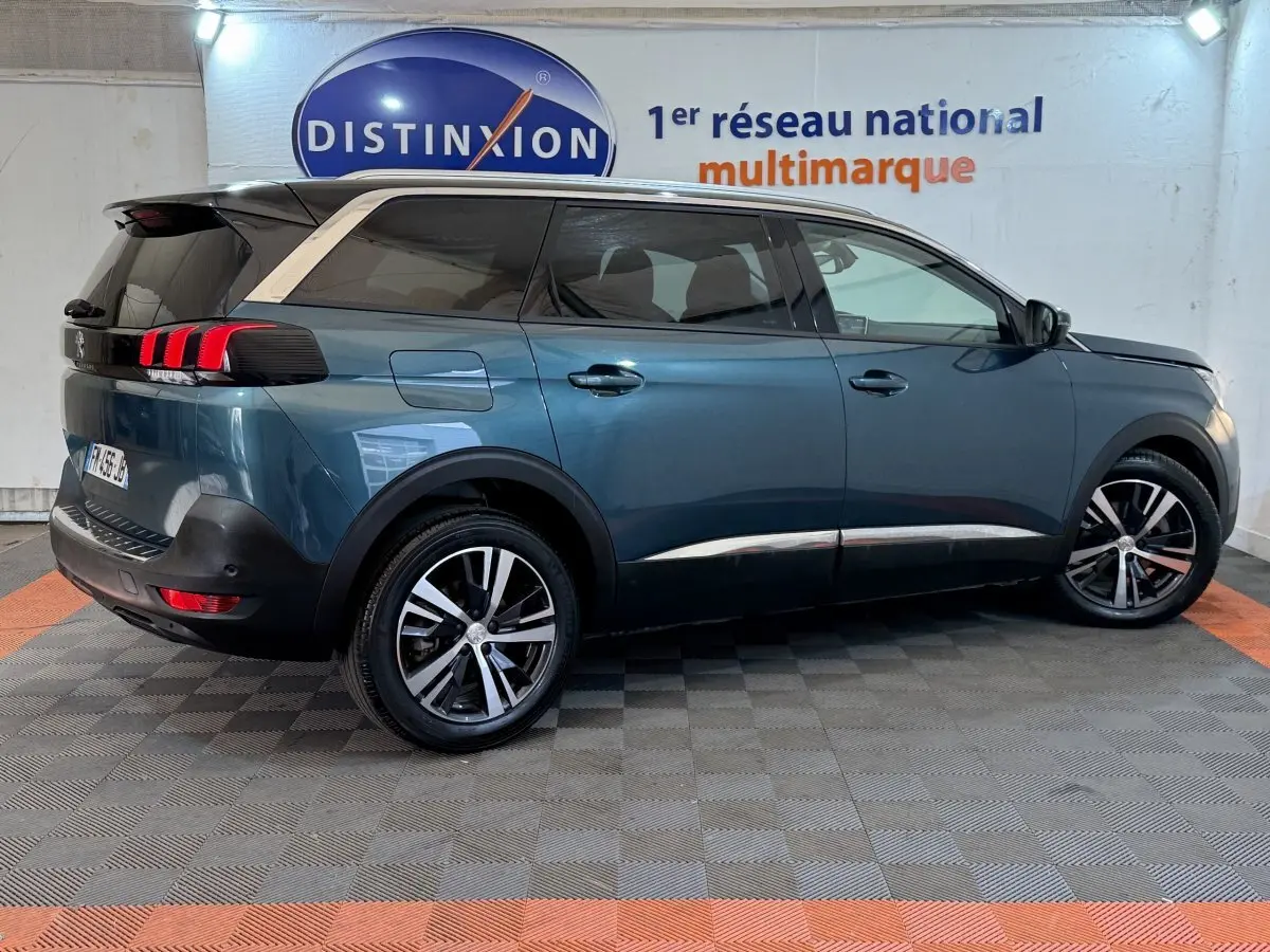 Vue latérale droite d’un Peugeot 5008 bleu métallisé avec toit noir et jantes alliage bicolores, en intérieur showroom.