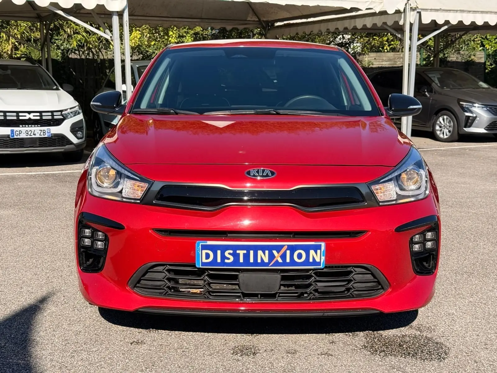 Vue frontale d'une KIA RIO rouge grenadine 2022 avec calandre noire et phares allumés, immatriculée Distinxion.