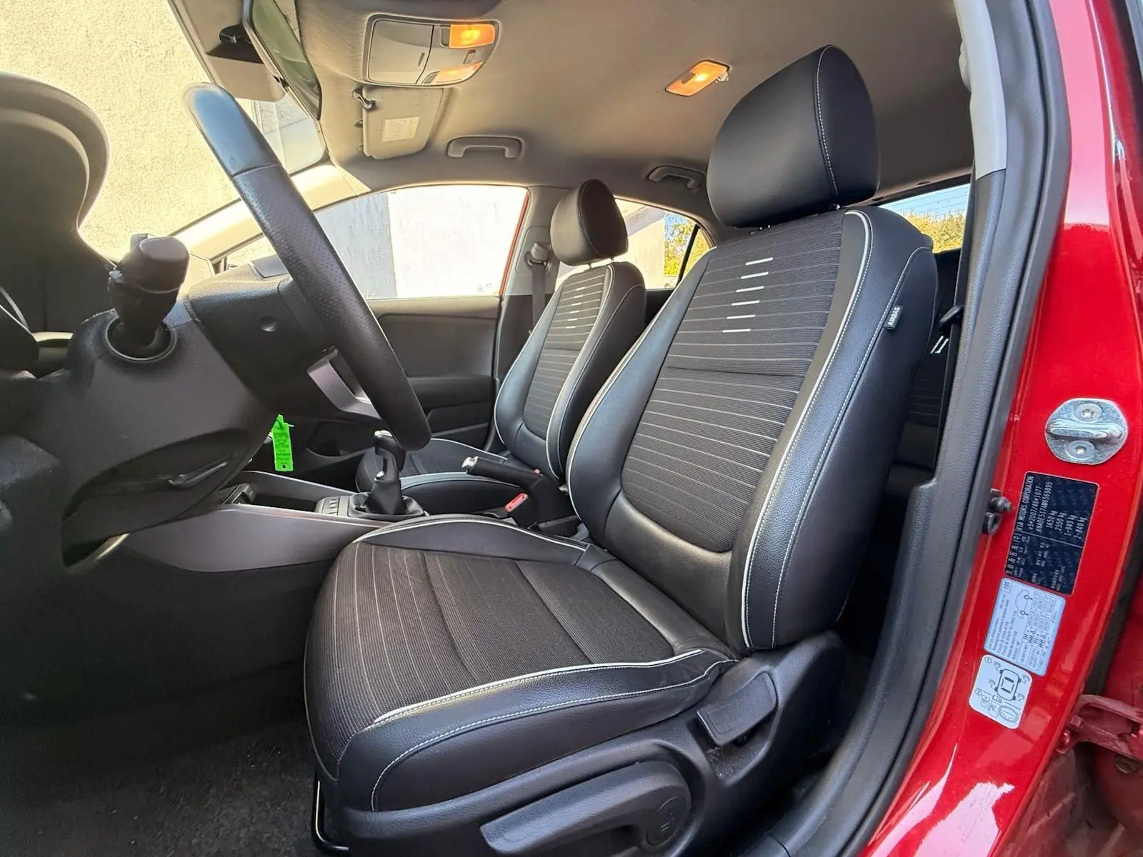 Intérieur côté conducteur d'une KIA Rio rouge grenadine 2022, sièges noirs avec surpiqûres blanches et volant visible.