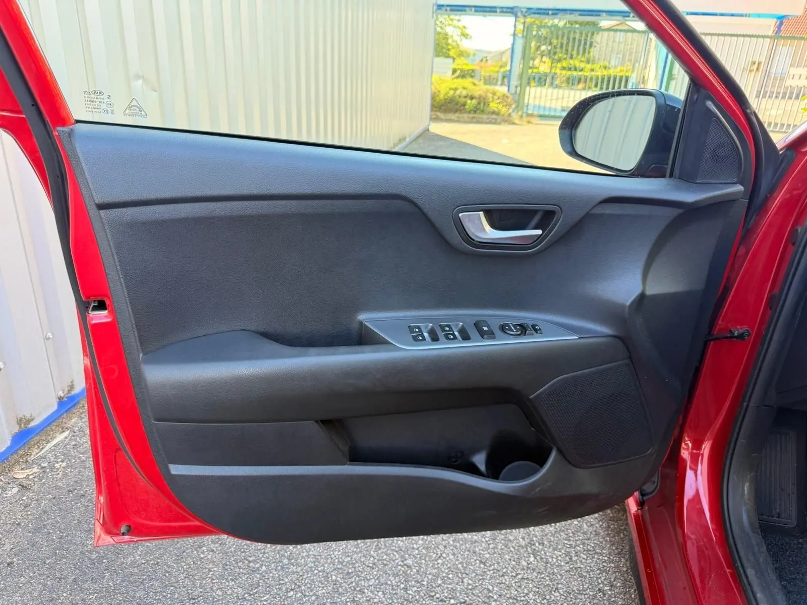 Porte avant gauche ouverte d'une KIA Rio rouge grenadine 2022, montrant les commandes électriques et le haut-parleur intégré.