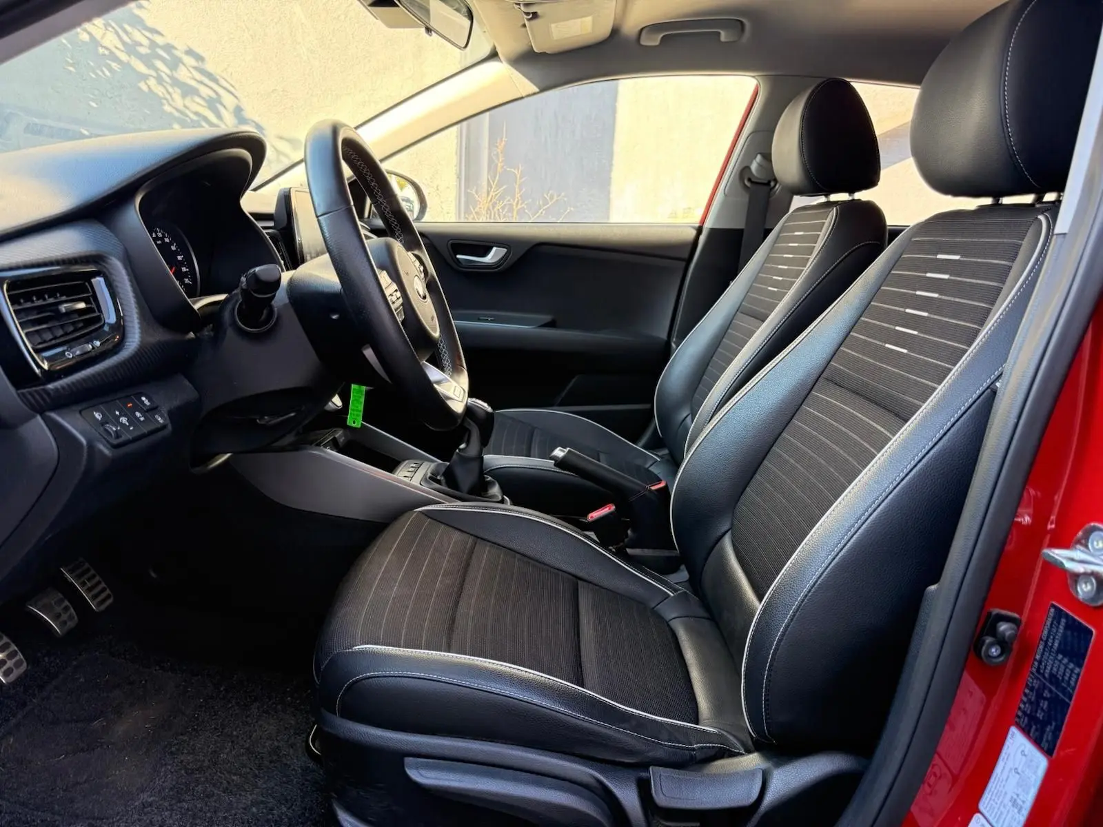 Intérieur côté conducteur d'une KIA RIO rouge grenadine 2022, sièges noirs avec surpiqûres blanches et volant multifonction.