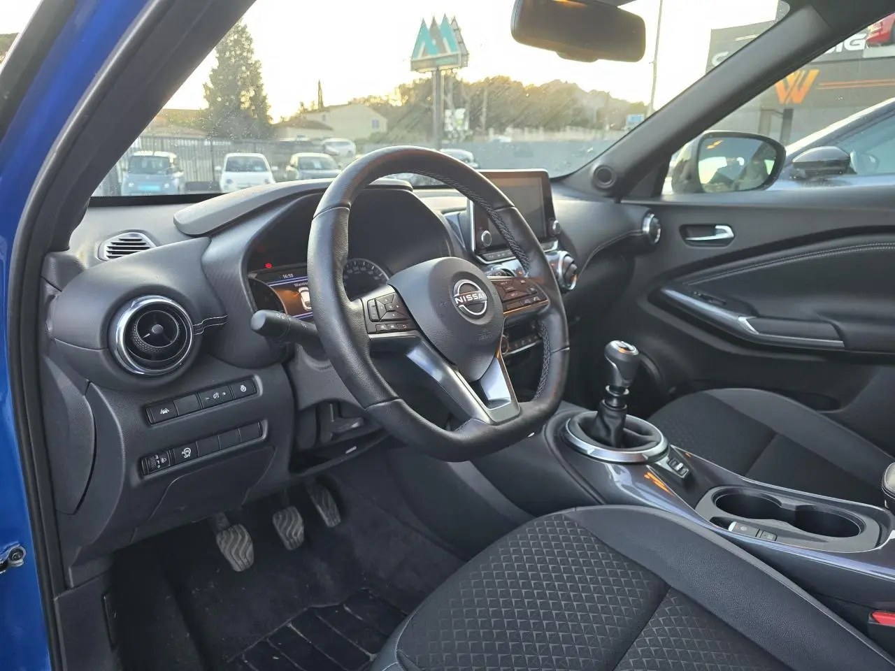 Intérieur avant droit du Nissan Juke II bleu magnetic 2024 avec volant cuir, boîte manuelle et console centrale moderne.
