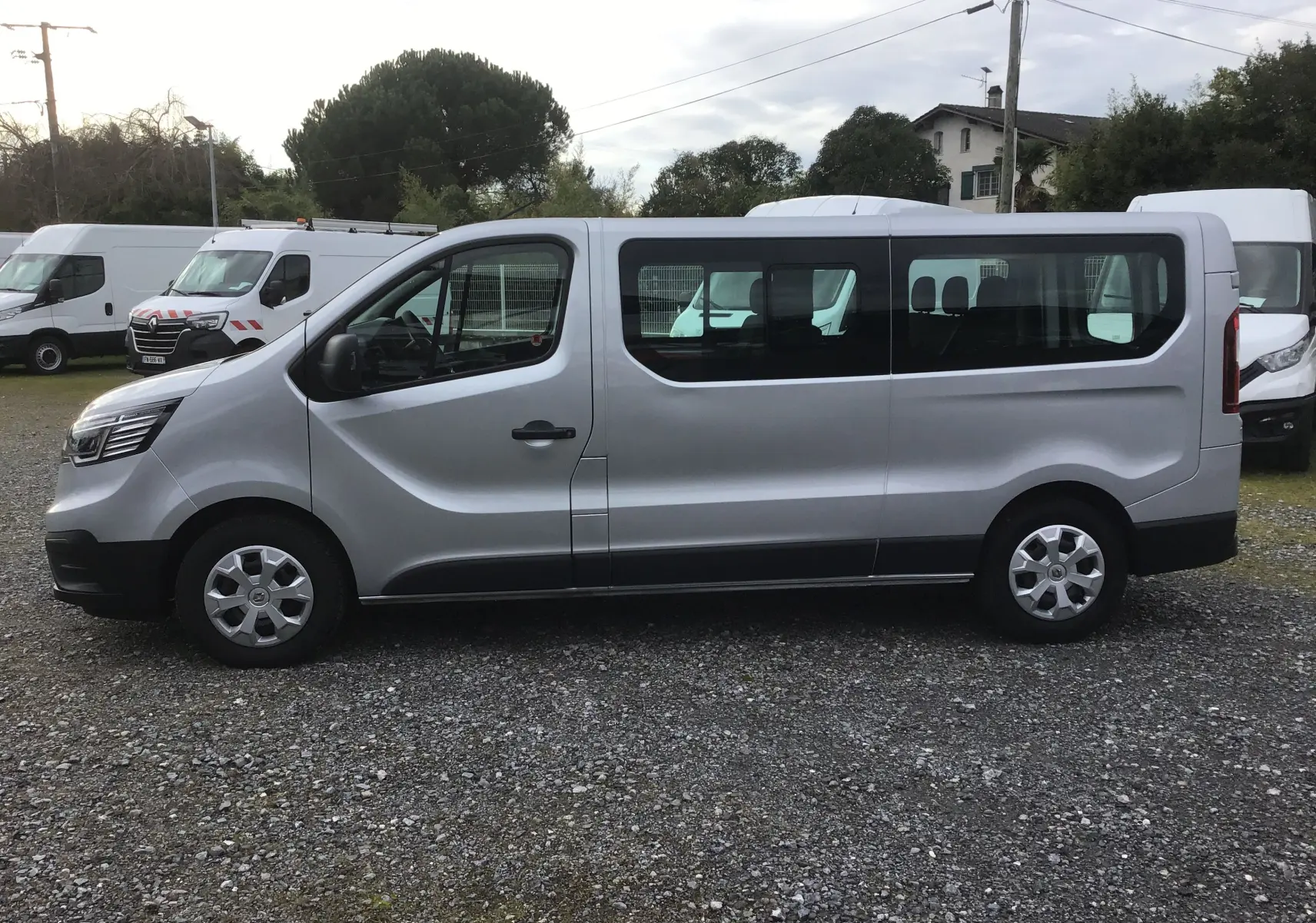 Vue de profil côté gauche d’un Renault Trafic Combi gris Highland 2022, grand fourgon 9 places stationné sur un sol gravillonné.