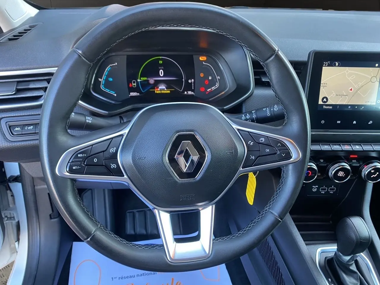 Vue rapprochée du volant en cuir noir avec logo Renault, tableau de bord numérique et écran tactile du Renault Clio V blanc.