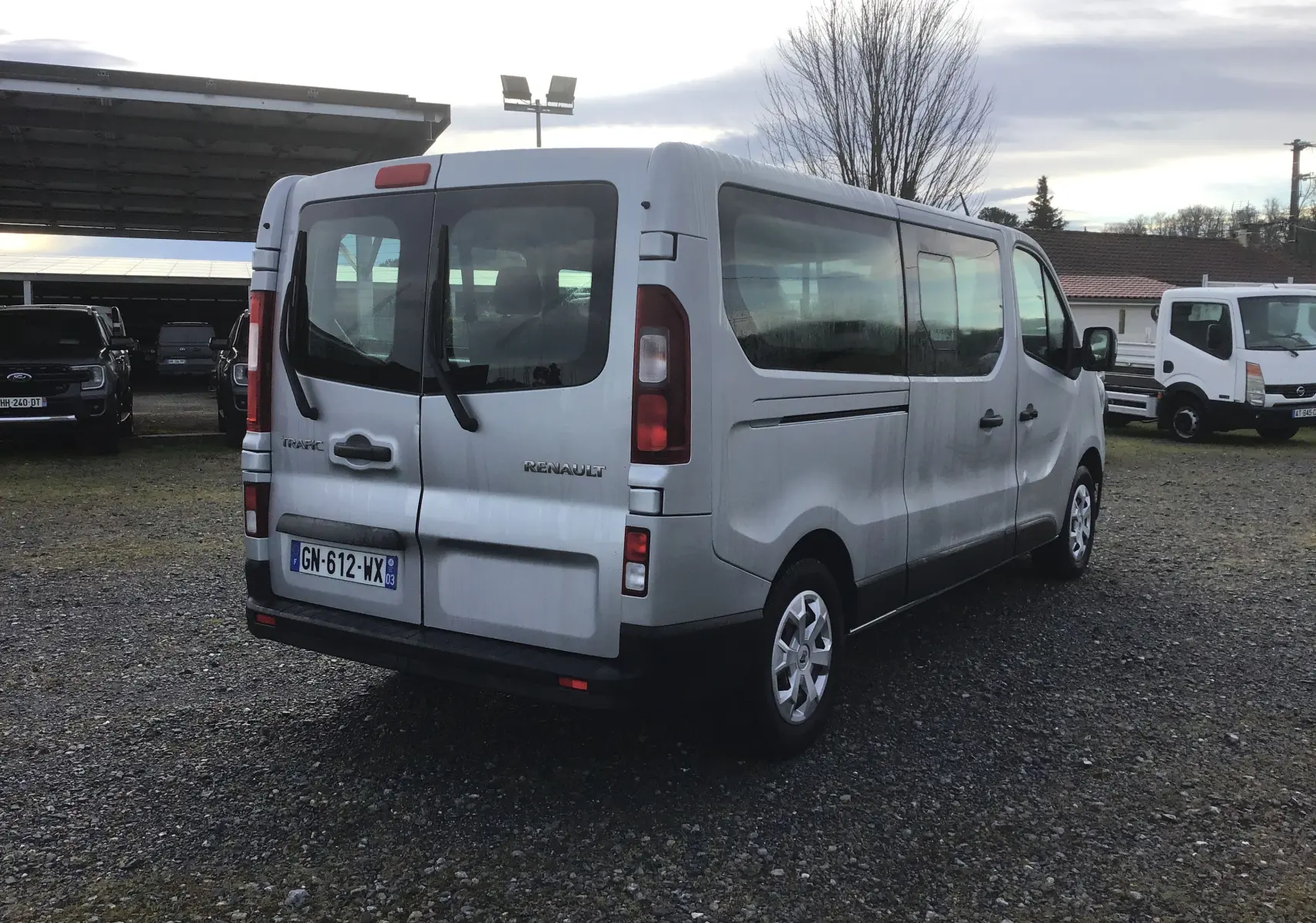 Vue 3/4 arrière droite d'un Renault Trafic Combi gris clair métal avec portes arrière vitrées sur un parking extérieur.