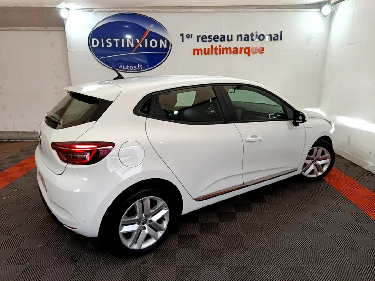 Vue 3/4 arrière droite d'une Renault Clio Business blanche, avec jantes alliage et feux arrière LED visibles en intérieur showroom.