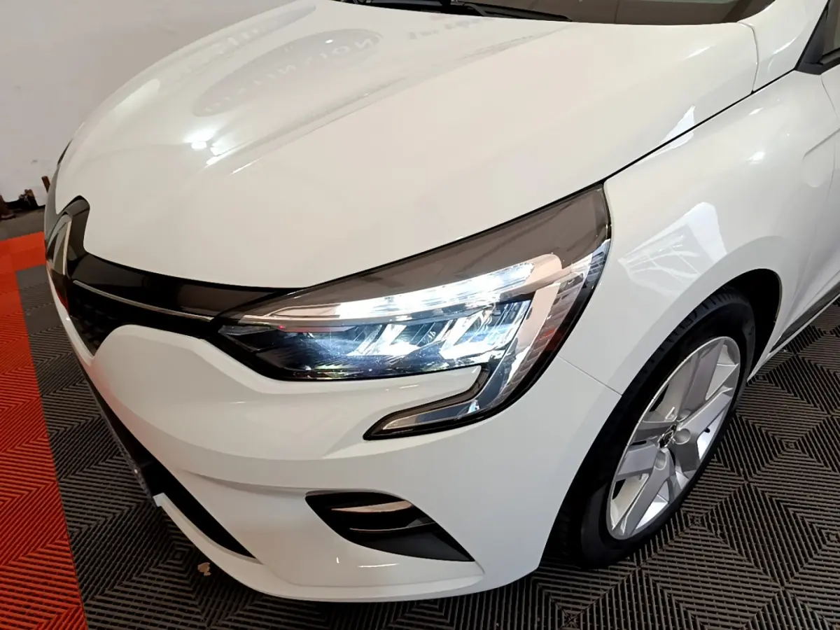 Vue 3/4 avant droit d'une Renault Clio Business blanche avec phares LED allumés et jantes alliage visibles.
