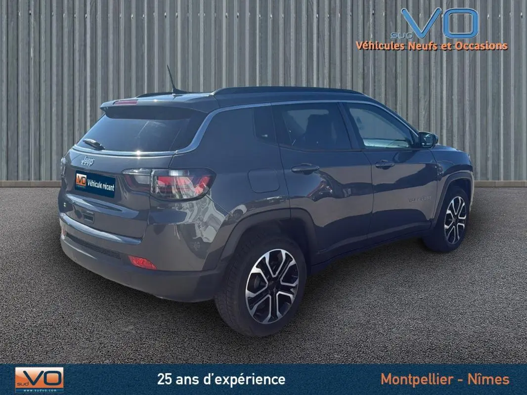 Jeep Compass gris 3/4 arrière droit, avec jantes 18 pouces et barres de toit visibles sur fond urbain.