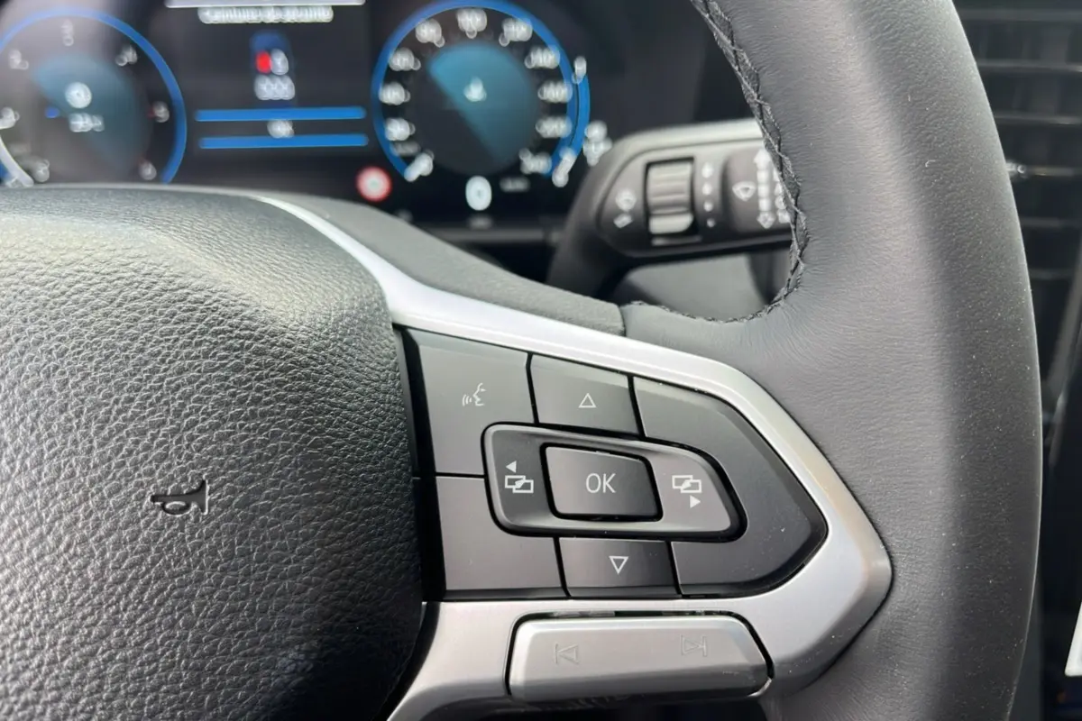 Gros plan sur le volant cuir multifonctions du Volkswagen Amarok noir, avec commandes intégrées et tableau de bord numérique flou en arrière-plan.