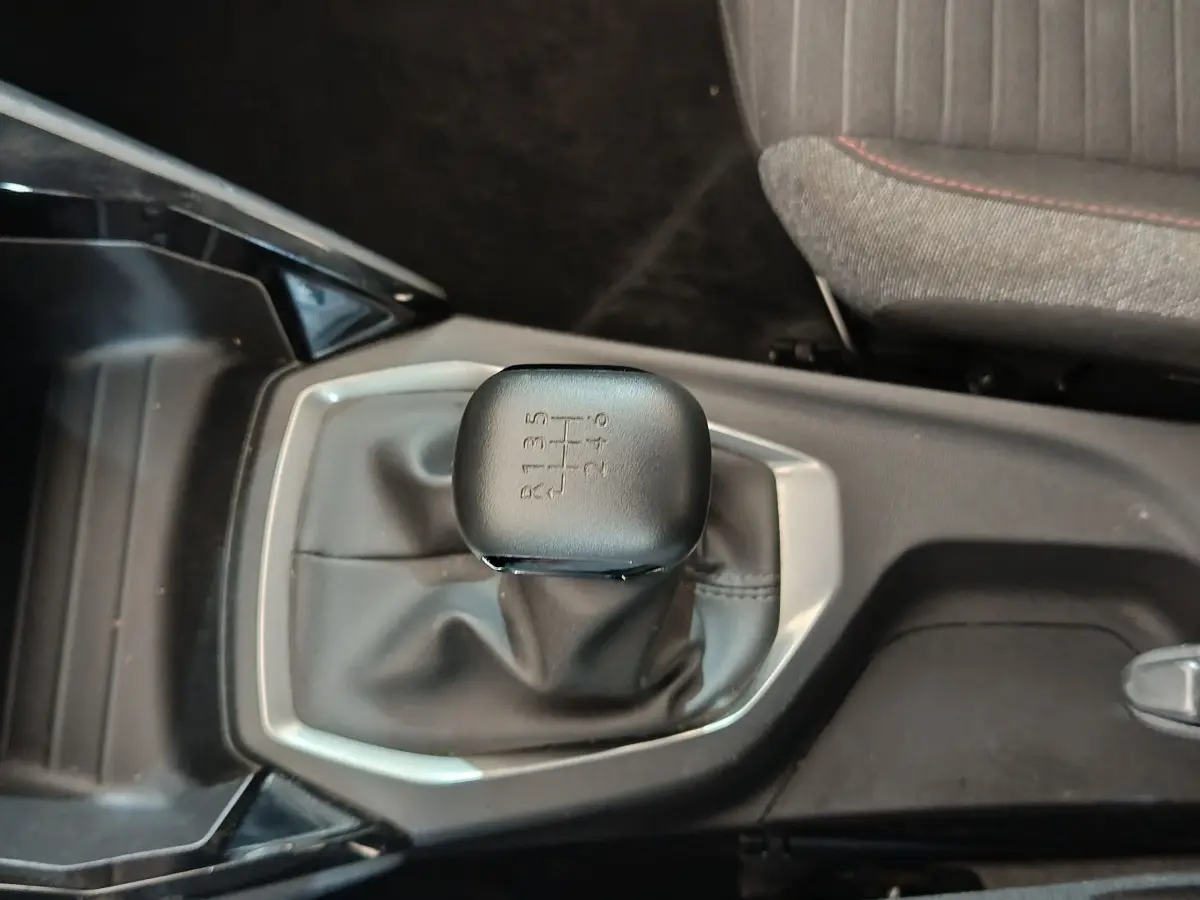 Le levier de vitesses manuel noir de la Peugeot 208 PureTech 100 vue de dessus, avec coutures grises et siège tissu adjacent.