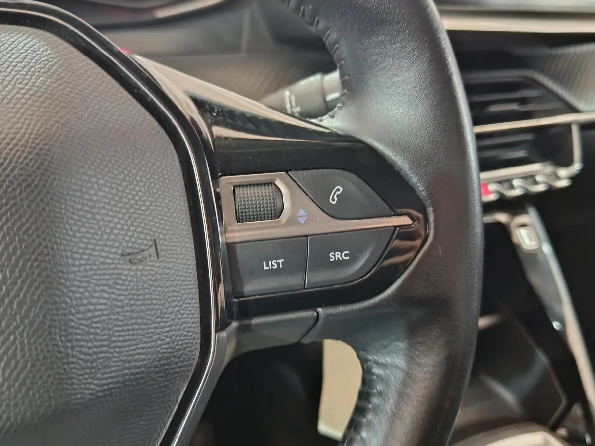 Gros plan sur les commandes gauche du volant cuir noir du Peugeot 208 blanc, avec boutons de téléphone et audio visibles.