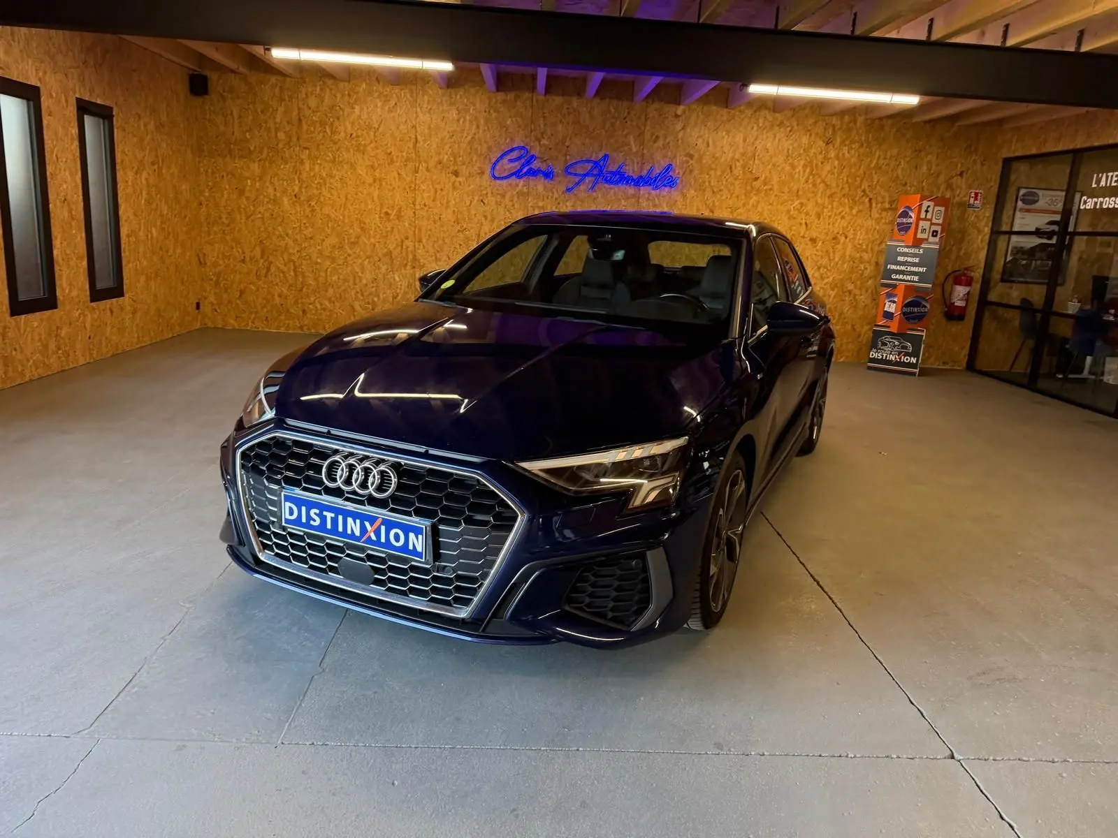 Audi A3 2.0 TDI S-Line bleu vue 3/4 avant droit dans un showroom avec mur en bois et enseigne lumineuse.