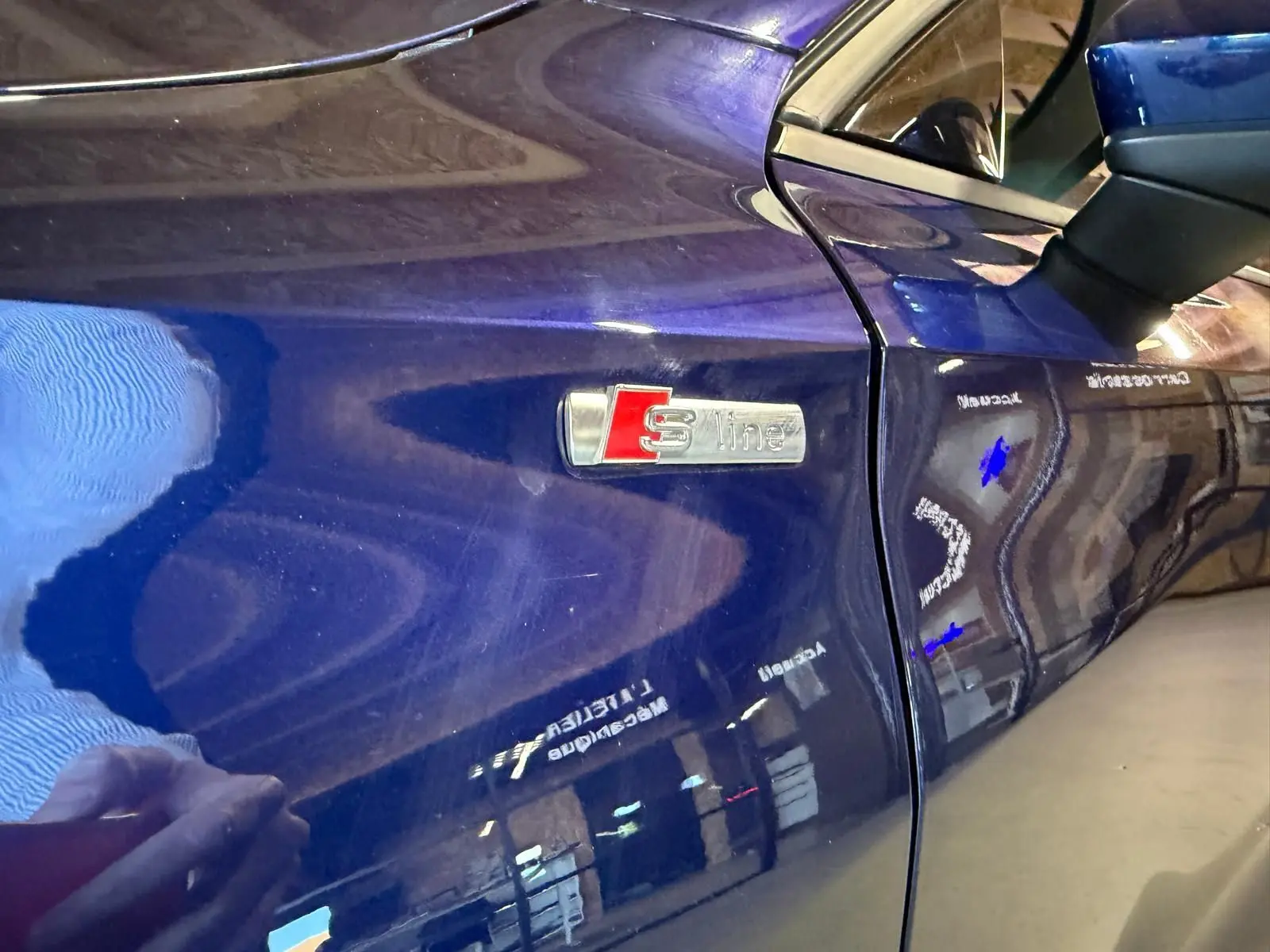 Gros plan sur l'aile avant gauche bleu foncé d'une Audi A3 S-Line, mettant en valeur l'emblème S-Line chromé et rouge.