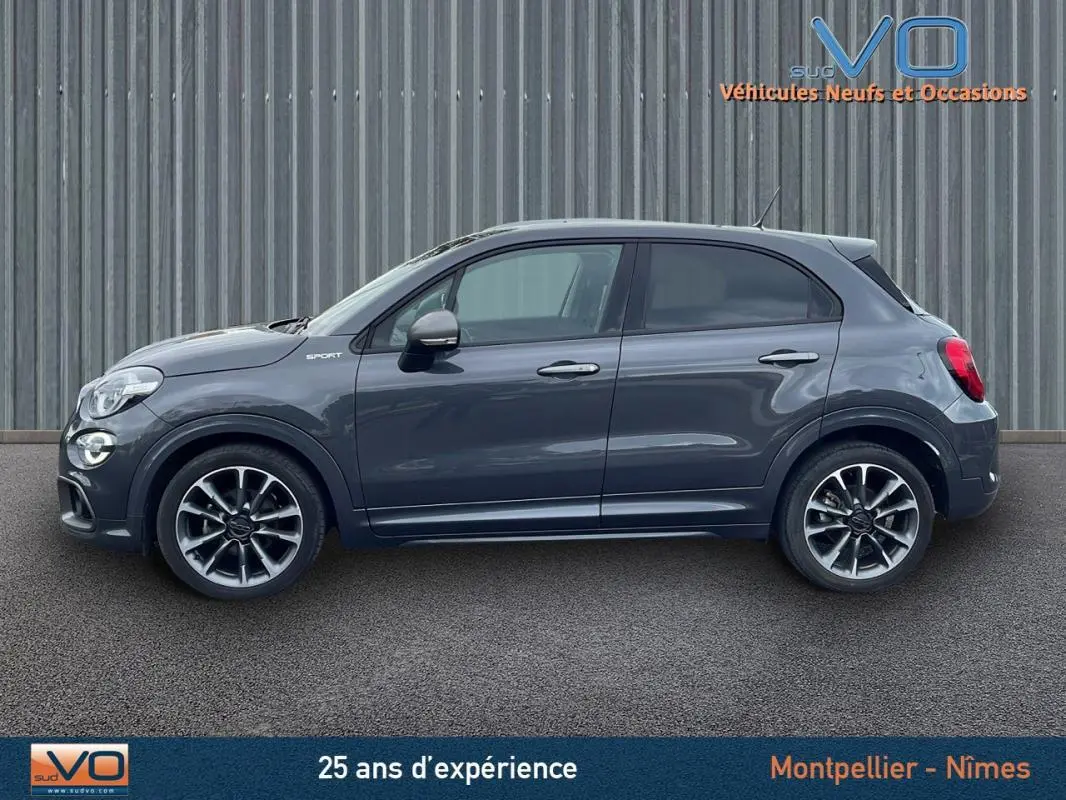 Profil côté gauche d'une FIAT 500X gris métallisé MY22 Sport avec jantes alliage 18 pouces sur fond industriel.