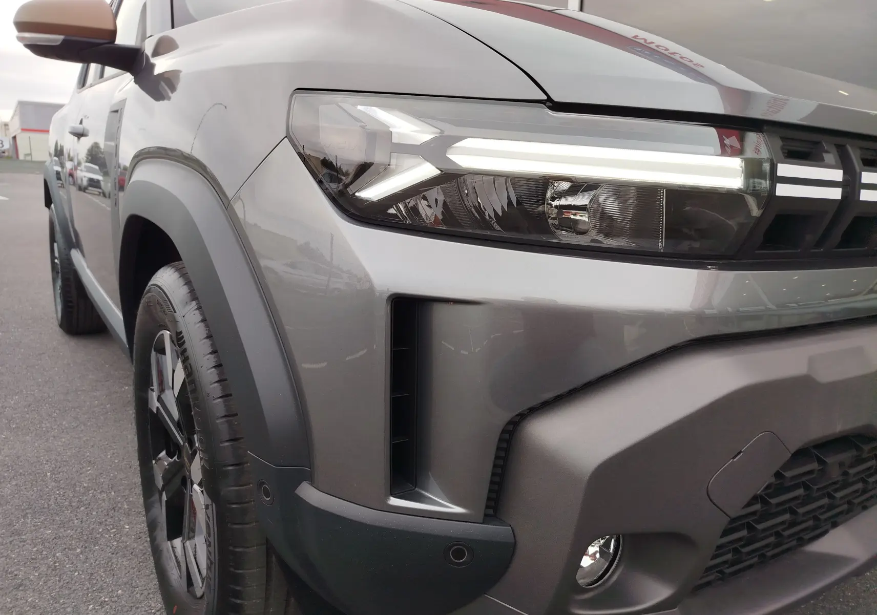 Gros plan sur l'avant droit gris schiste du Dacia Duster 2025, mettant en valeur son phare LED et son bouclier robuste.