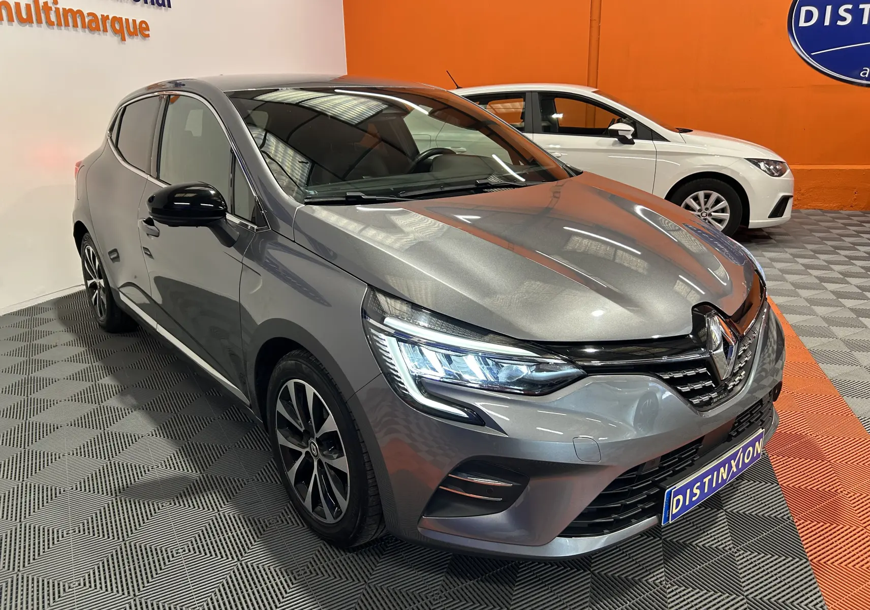 Vue 3/4 avant gauche d'une Renault Clio V gris métallisé 2023 avec phares LED allumés et caméra 360° visible sur le pare-chocs.