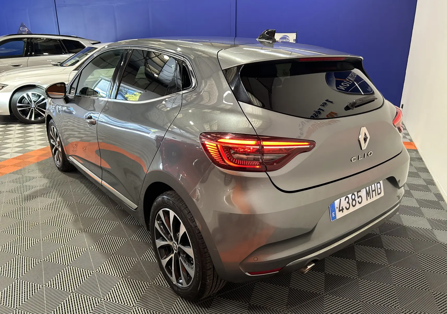 Vue 3/4 arrière droite d'une Renault Clio V TCe 90 TECHNO gris métallisé avec feux arrière allumés en intérieur showroom.