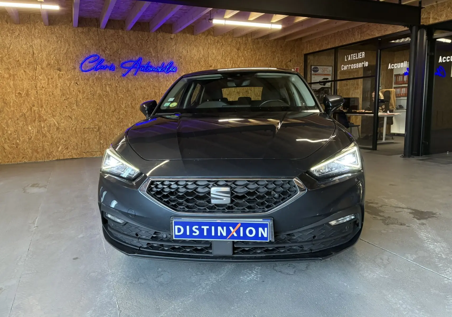 Vue de face d'une SEAT Leon gris foncé 2021 avec phares allumés et plaque Distinxion dans un showroom.