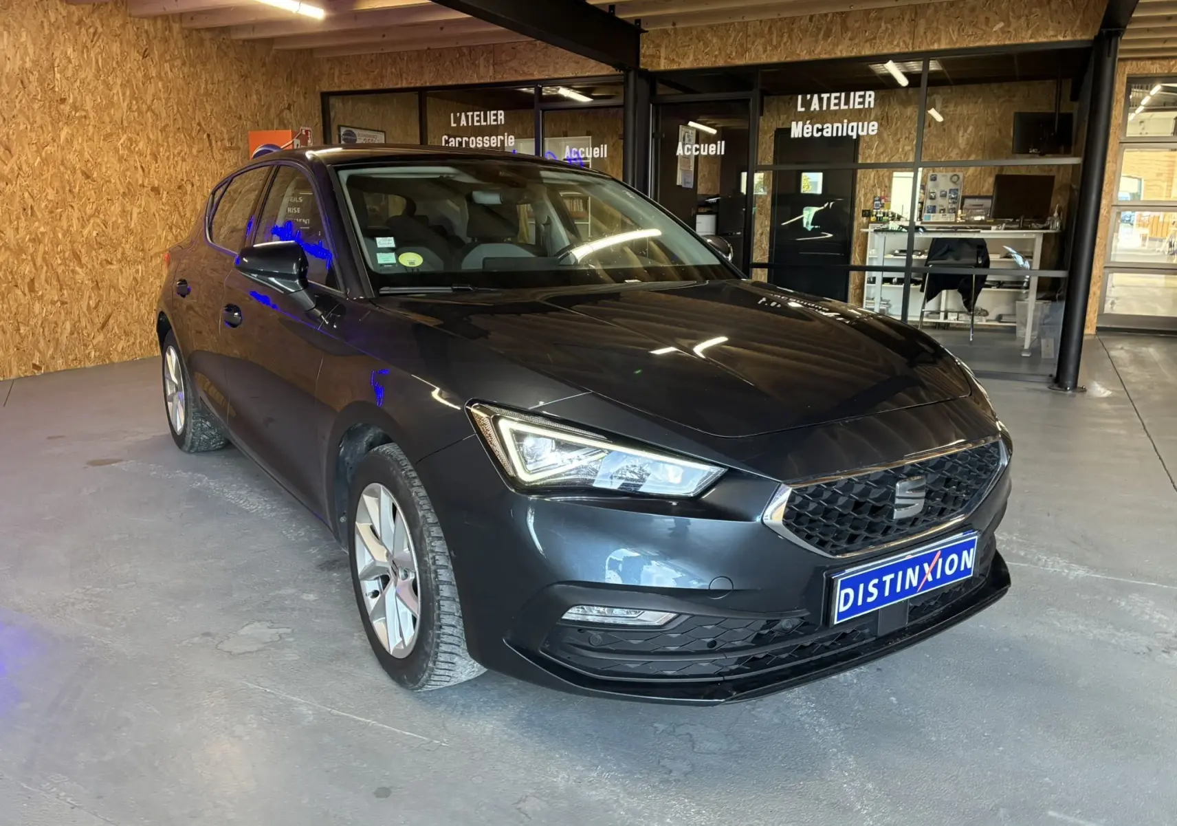 SEAT Leon gris 3/4 avant droit, phare LED allumé, calandre noire et intérieur d'atelier en arrière-plan.