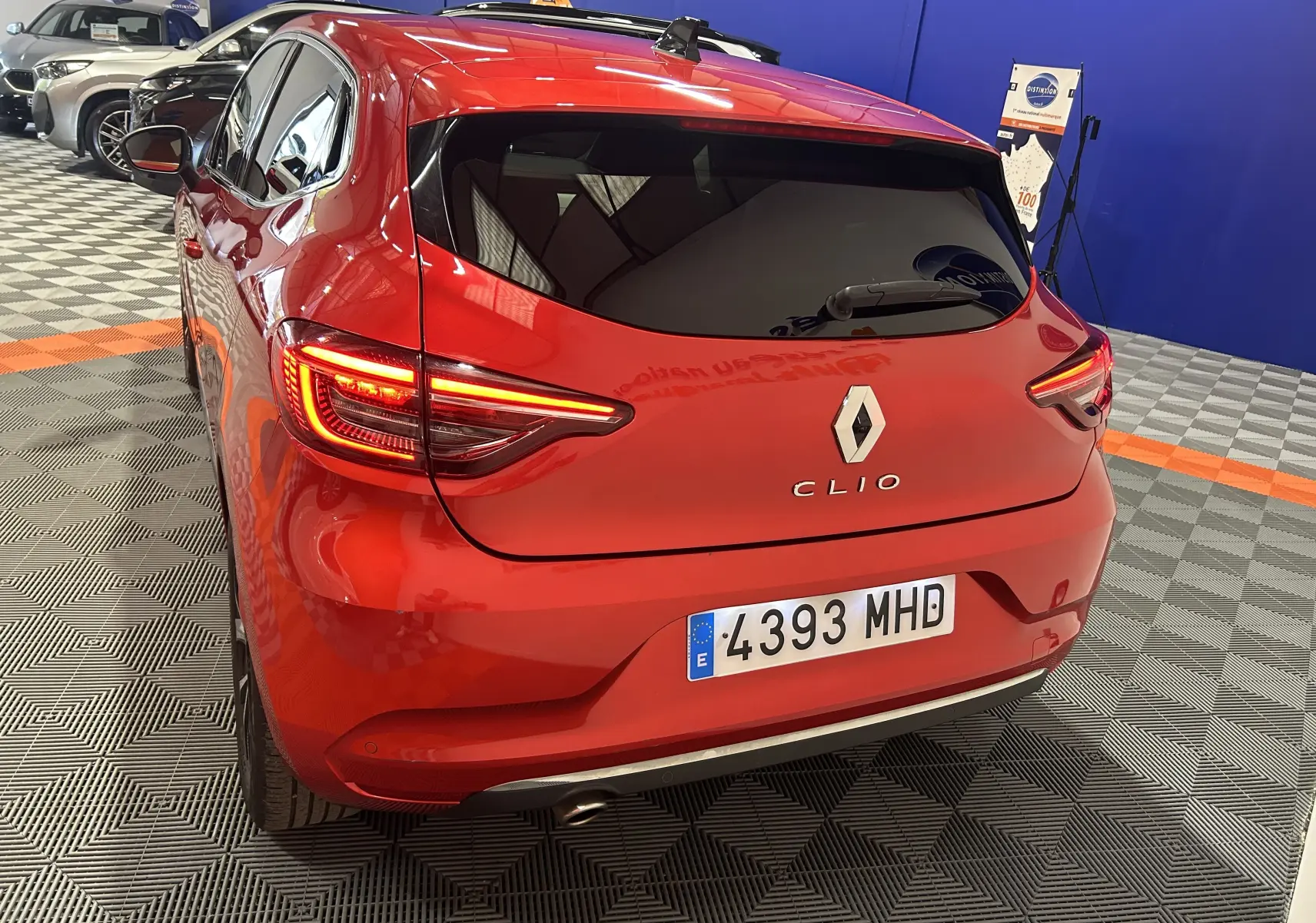 Vue 3/4 arrière d'une Renault Clio V rouge 2023 avec feux allumés dans un showroom intérieur.
