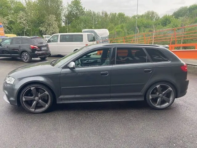 Profil côté gauche d'une Audi RS3 Sportback gris Daytona avec jantes noires et vitres teintées, stationnée sur sol humide.