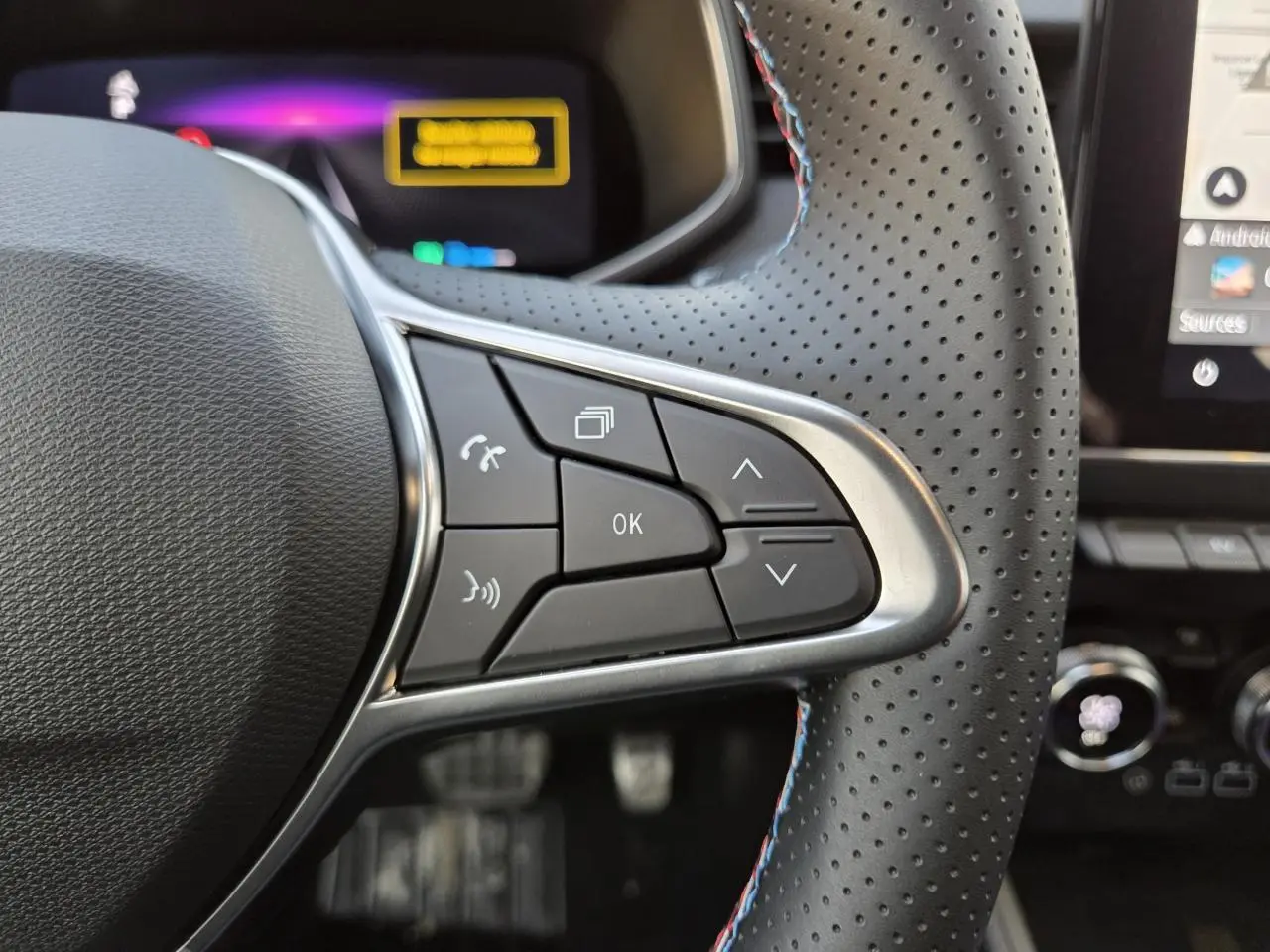 Gros plan sur les commandes du volant perforé avec surpiqûres tricolores de la Renault Clio V hybride Gris Rafale.