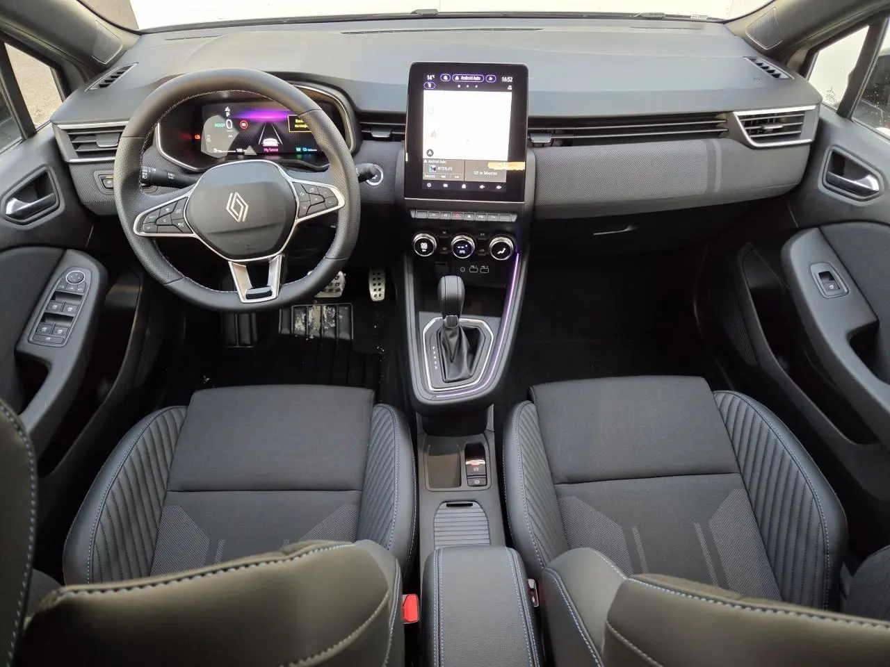 Vue intérieure avant de la Renault Clio V hybride 2025, sièges noirs avec surpiqûres bleues et écran tactile vertical.