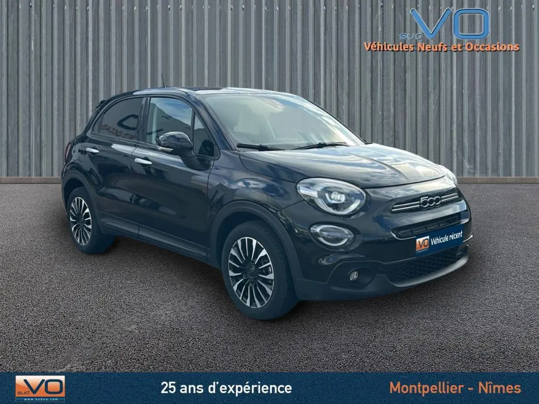 Vue 3/4 avant d'une FIAT 500X noire avec jantes classiques 16 pouces sur un sol goudronné devant un mur métallique.