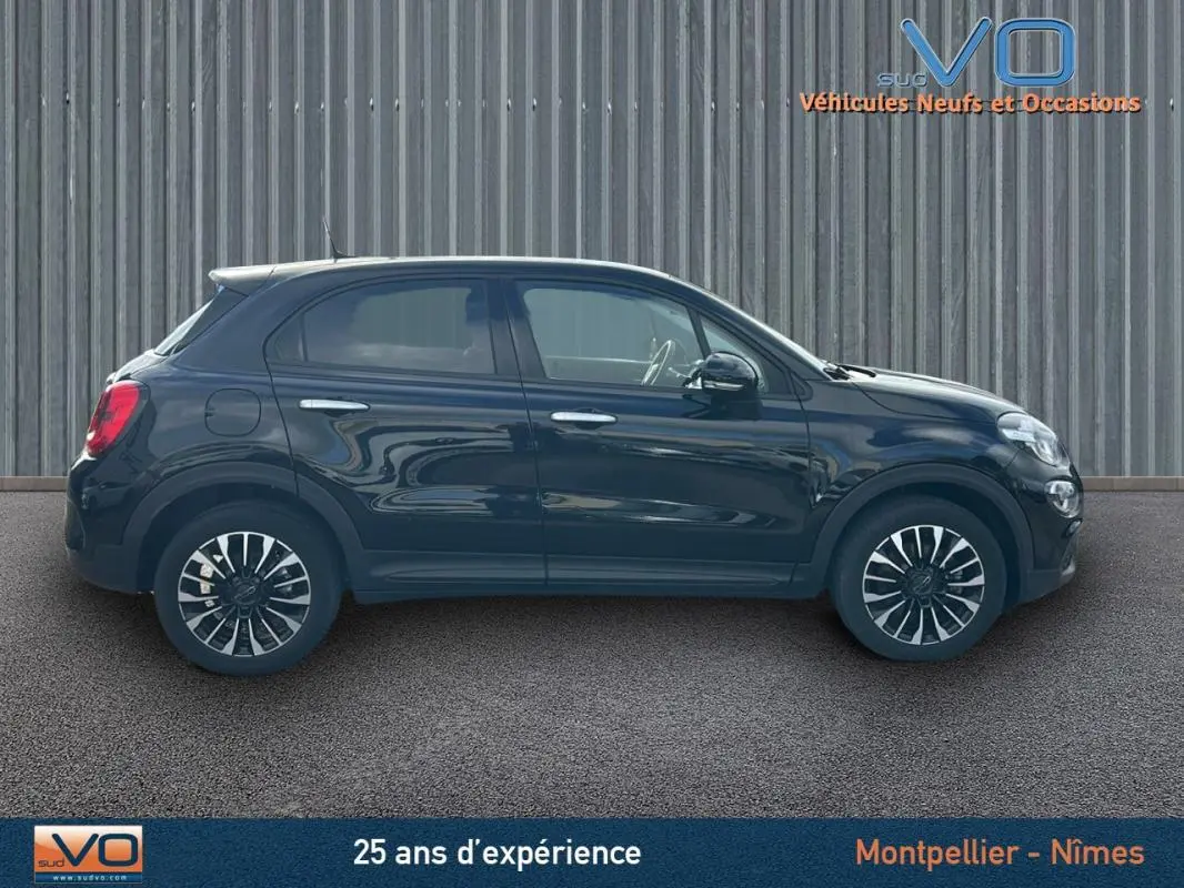 Profil droit d'une FIAT 500X noire 2023 avec jantes classiques 16 pouces, stationnée devant un mur métallique.