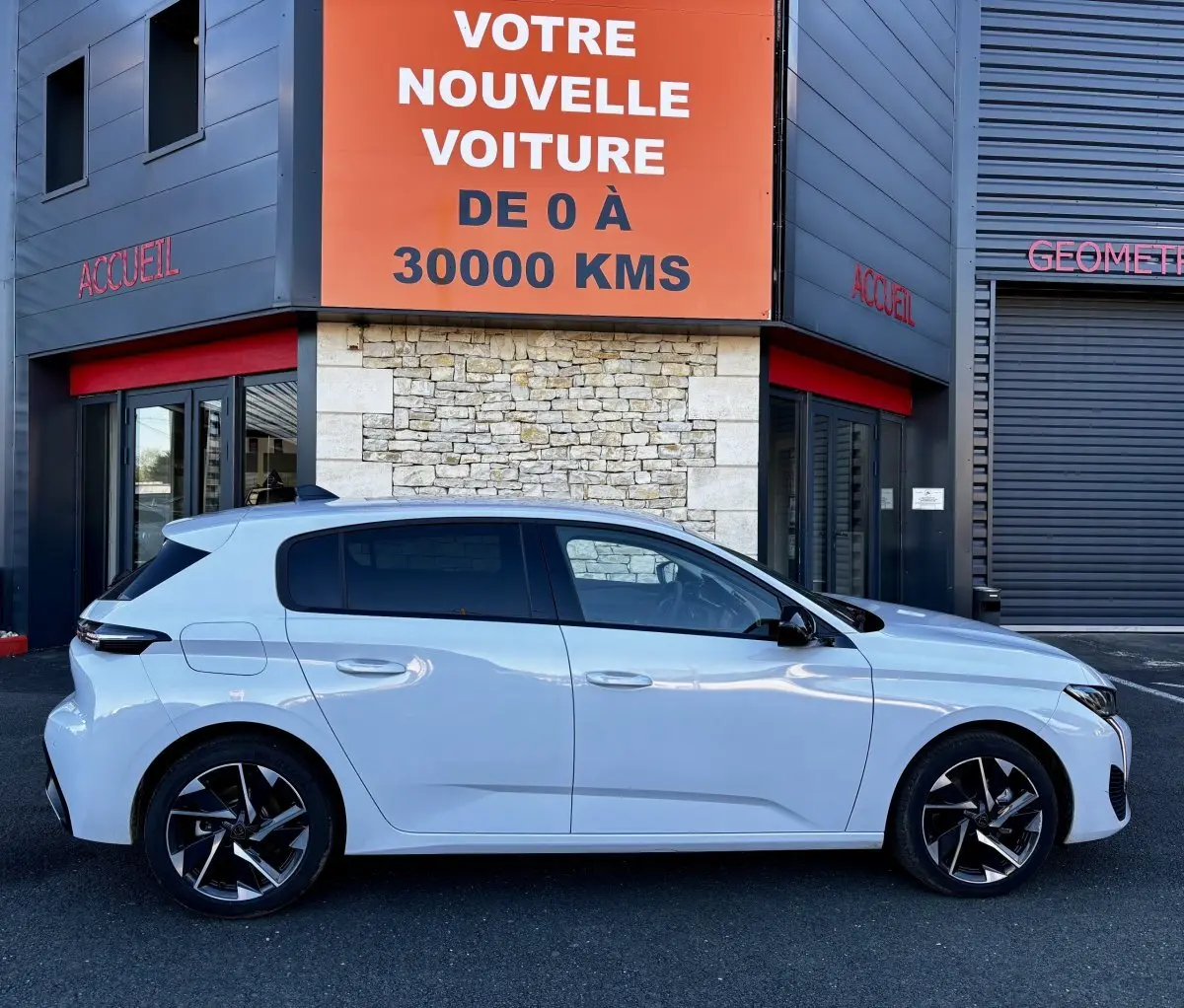 Vue latérale droite d'une Peugeot 308 Hybrid 145 e-DCS6 Allure blanche, garée devant un bâtiment avec panneau orange.
