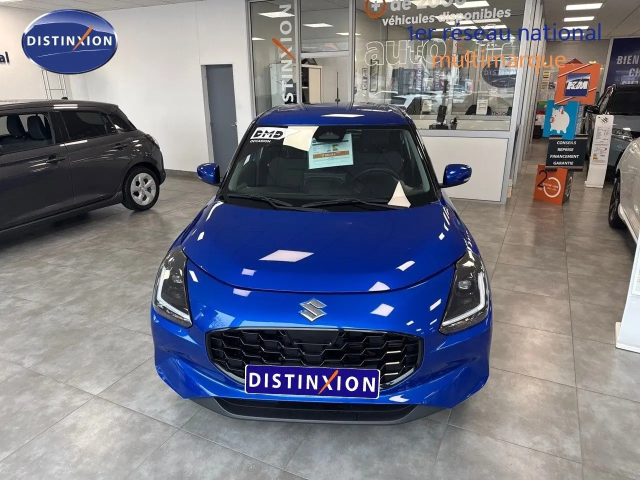 Vue avant d'une Suzuki Swift 1.2 Pack Hybrid bleu brillant exposée en showroom avec calandre noire et phares LED.