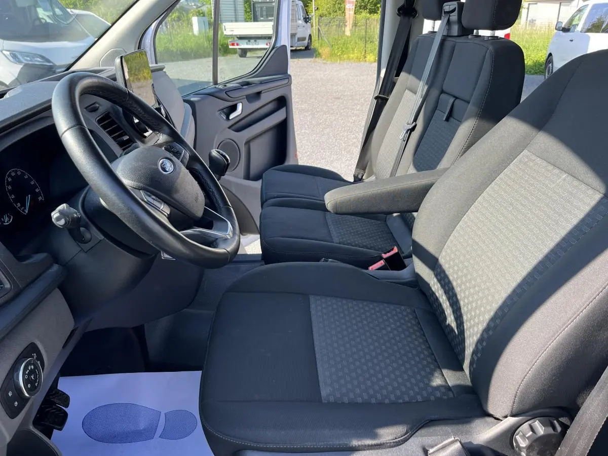 Intérieur du Ford Transit Custom blanc 2021, vue côté conducteur montrant les sièges avant et le volant cuir.