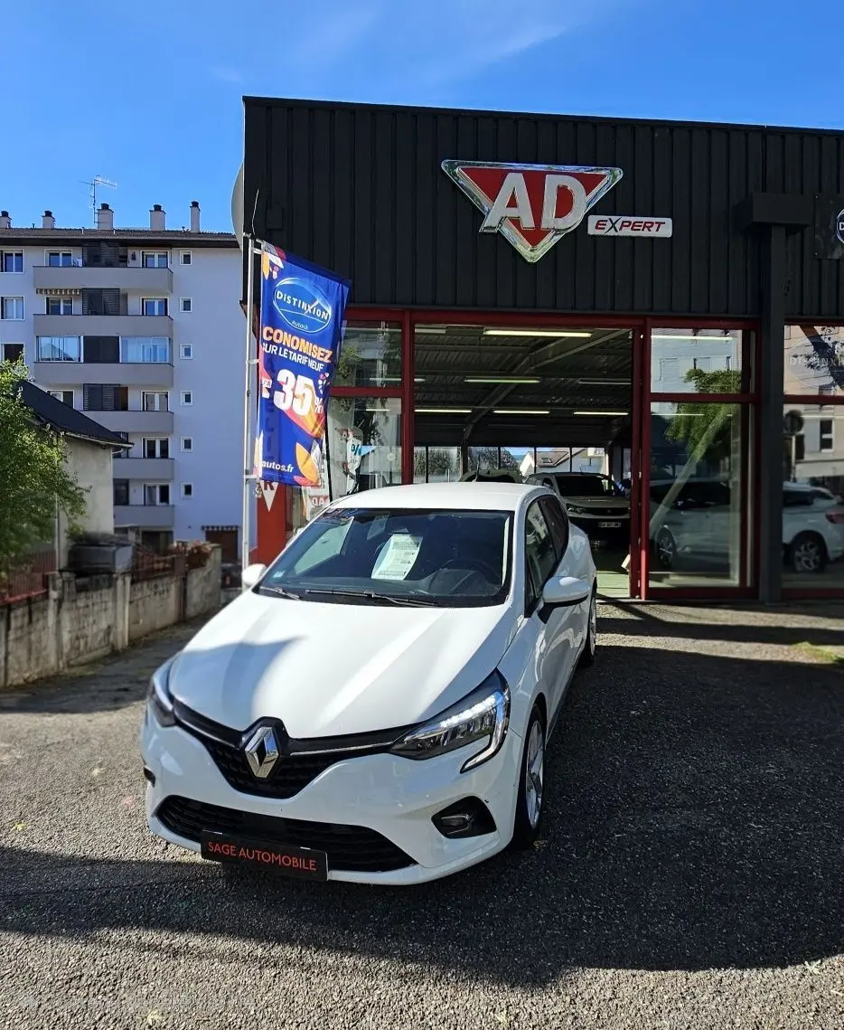 Renault Clio blanc glacier vue 3/4 avant droit devant un garage AD Expert avec une affiche promotionnelle bleue.