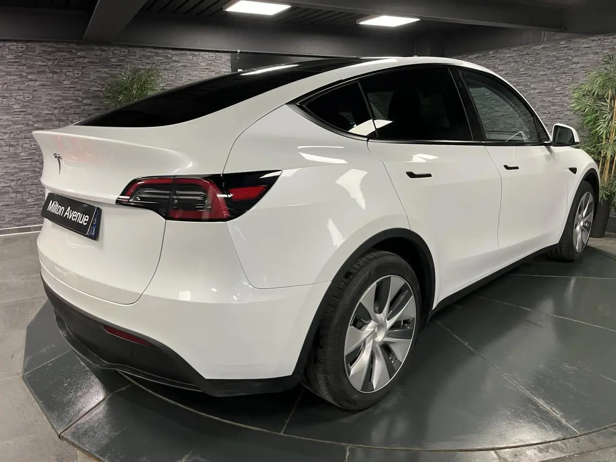Tesla Model Y blanc nacré vu en 3/4 arrière droit dans un showroom avec jantes alu et toit panoramique noir.