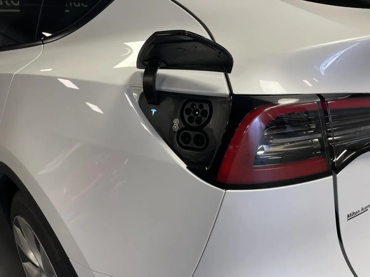 Prise de recharge ouverte côté gauche arrière d'une Tesla Model Y blanche avec feux arrière LED visibles.