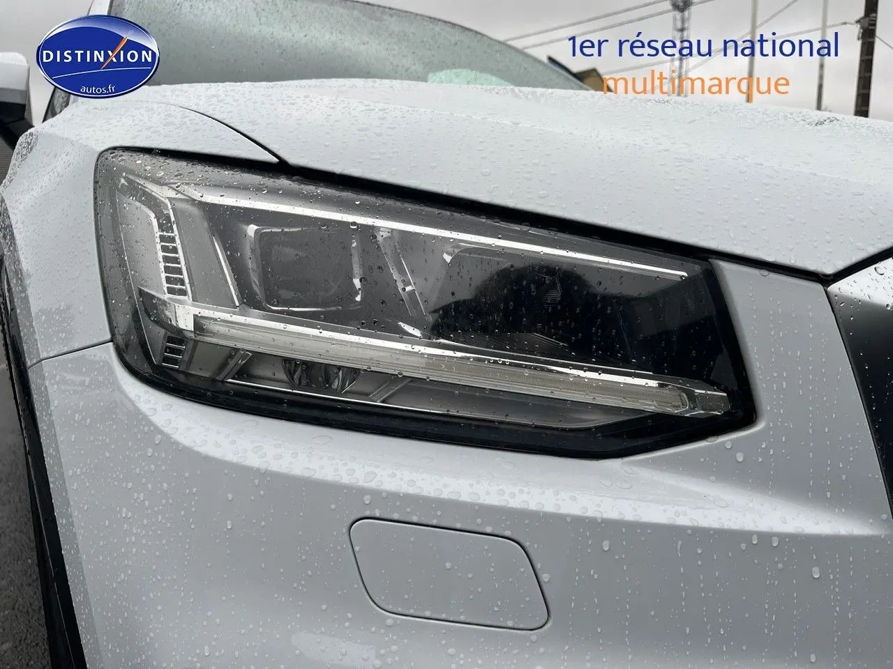 Gros plan sur le phare avant gauche d’un Audi Q2 blanc Ibis avec gouttes de pluie visibles sur la carrosserie.