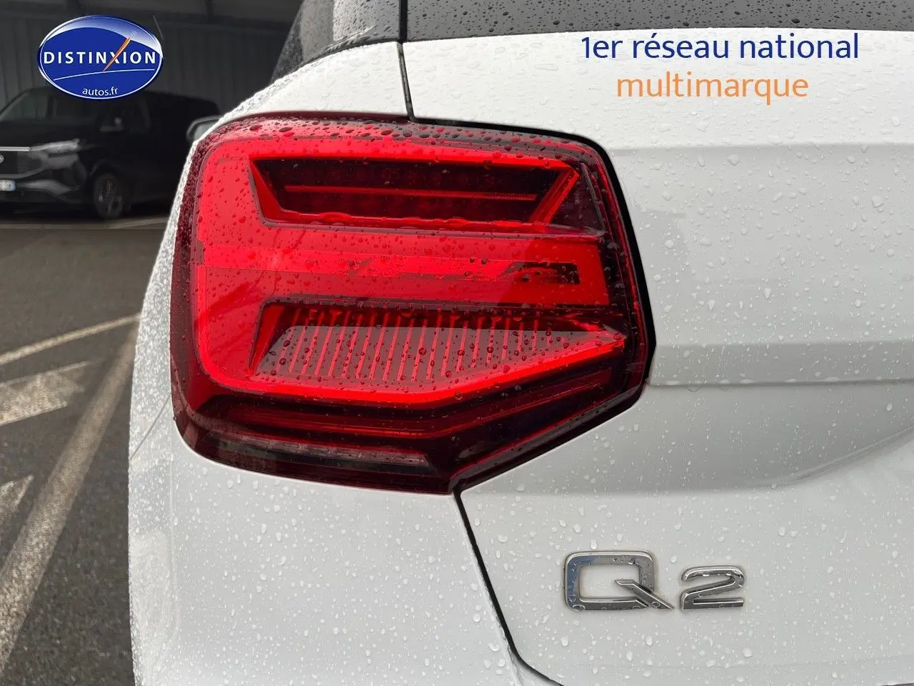 Gros plan sur le feu arrière gauche d’un Audi Q2 blanc Ibis avec logo Q2 visible sous la pluie.