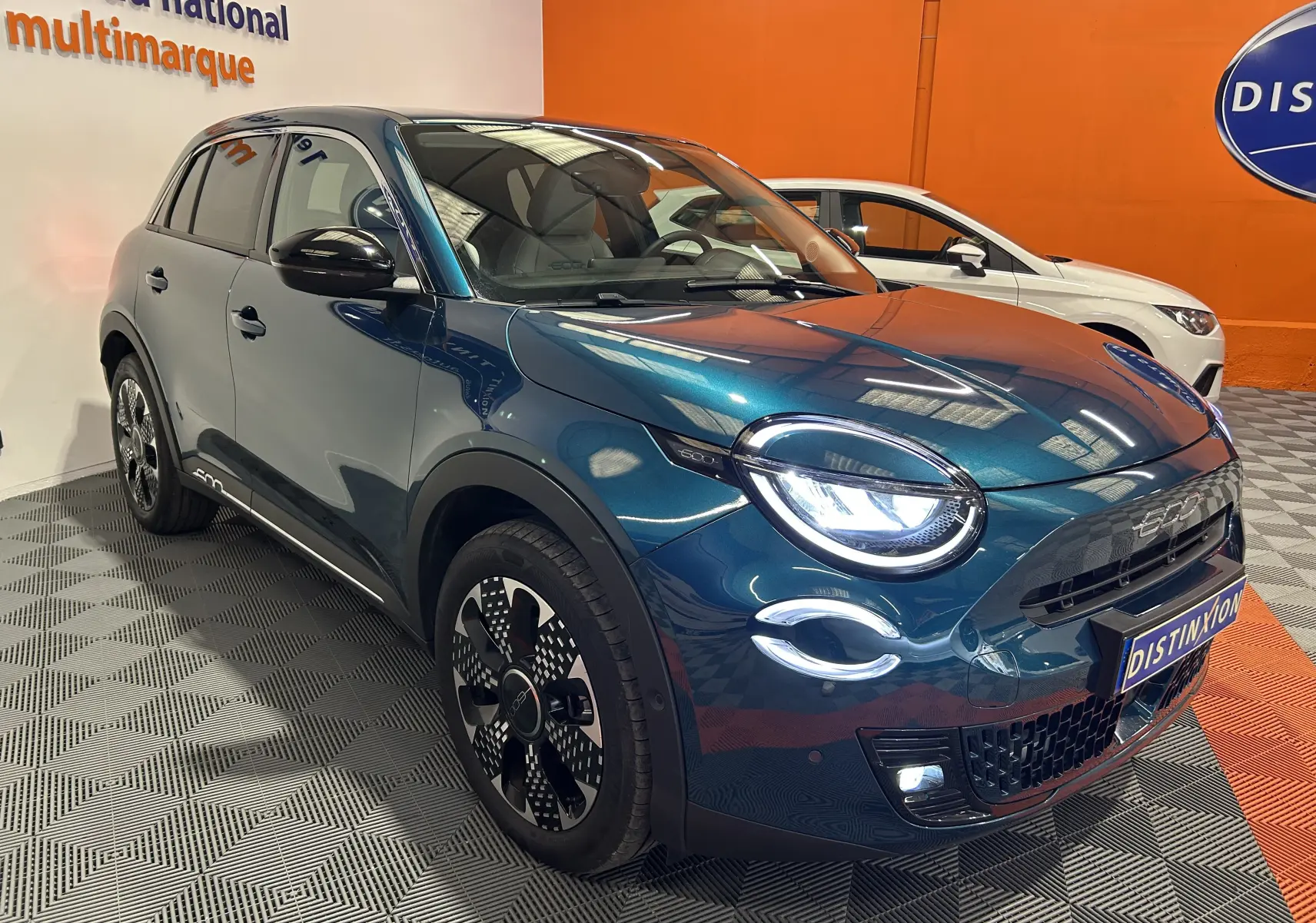 Vue 3/4 avant droite d'une FIAT 600 Hybrid 100 La Prima bleu métallisé avec phares LED allumés en showroom.